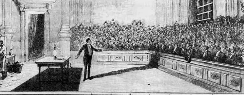 Black-and-white engraving of a man standing beside a table with apparatus, addressing a large seated audience in a columned hall.