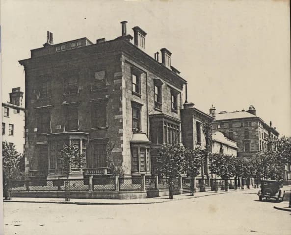 Black-and-white photograph of a three-story brownstone corner townhouse on a tree-lined city street, with a vintage automobile parked at the curb.