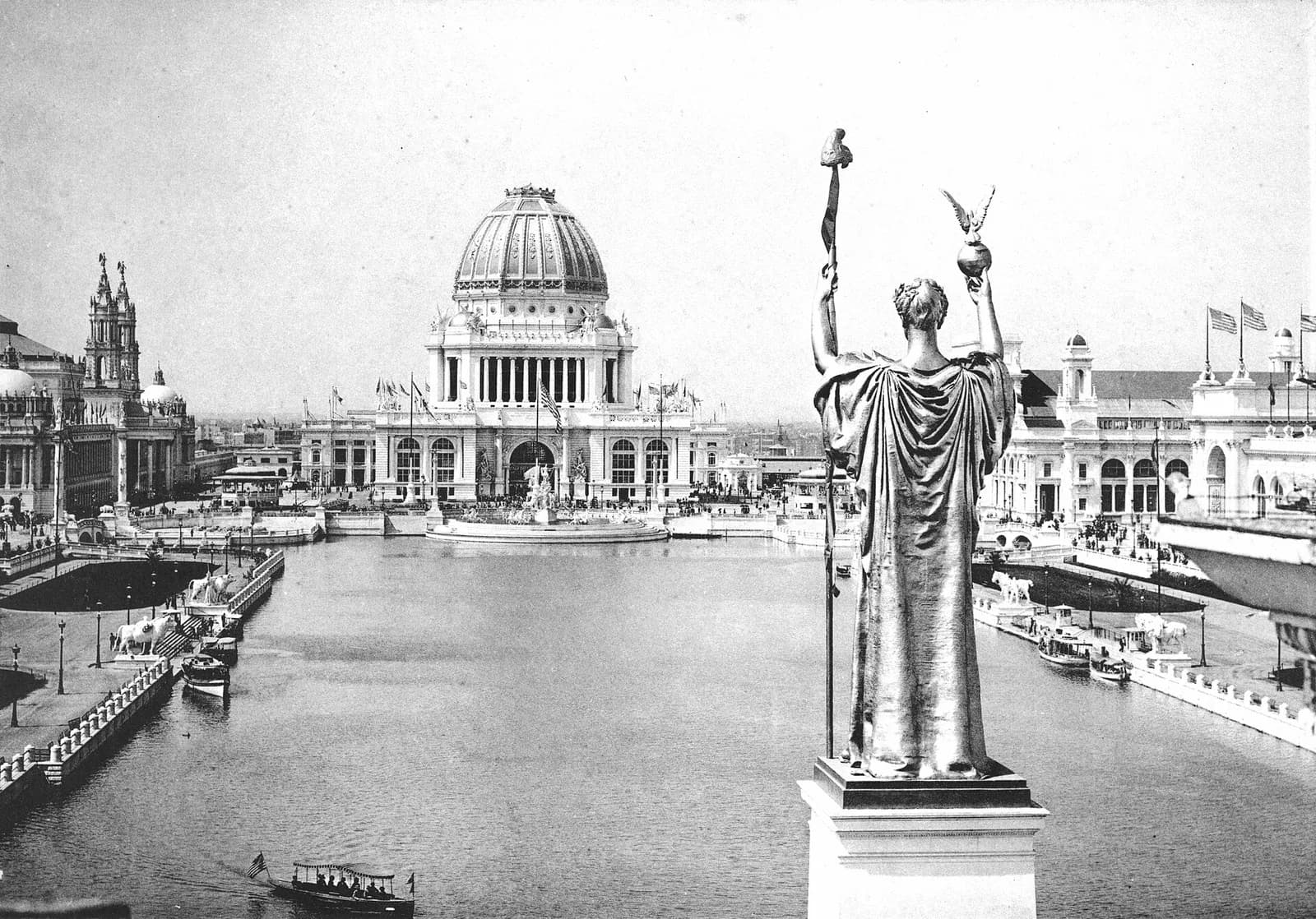 Black-and-white photograph of a large gilded statue overlooking a reflecting lagoon lined with domed neoclassical exposition buildings.