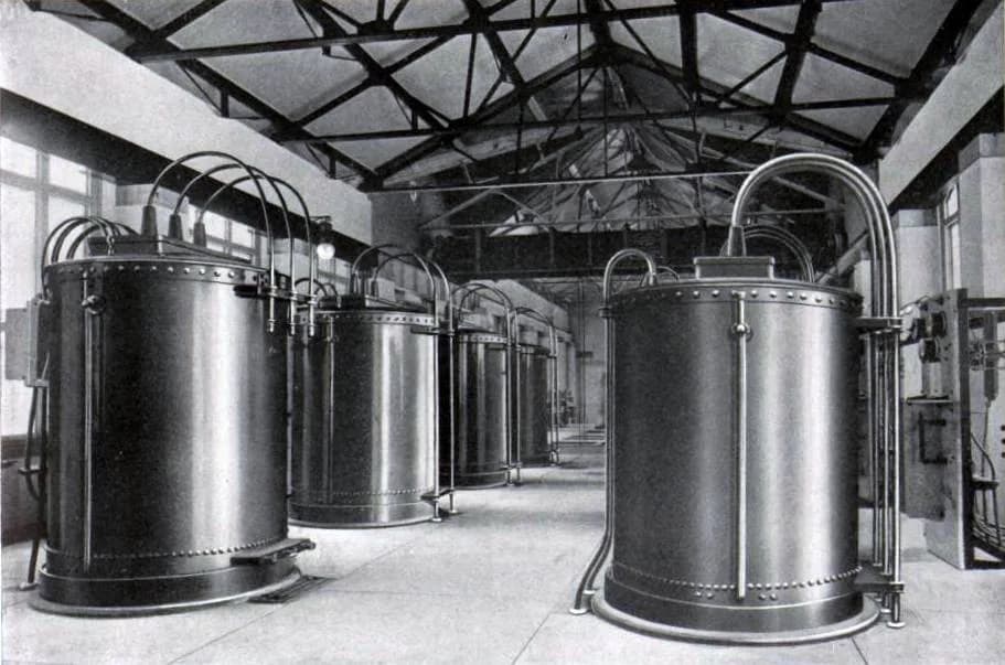 Black-and-white photograph of a row of large cylindrical metal transformers standing in an industrial hall with a high trussed ceiling.