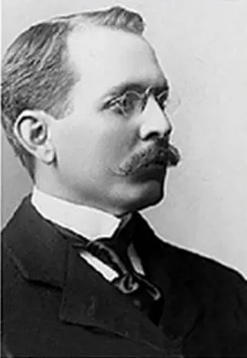 Black-and-white side-profile photograph of a young man with a mustache and round wire-rimmed glasses, wearing a high collar and dark suit.