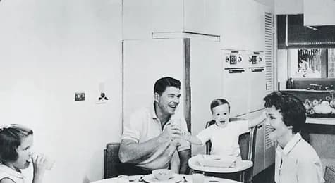 A black-and-white photograph of a man, a woman, and two children at a table in a kitchen with built-in double wall ovens.