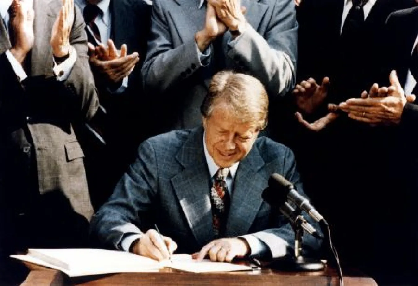 A photograph of a man seated at a desk signing a document, surrounded by a group of applauding people in suits, with a microphone in the foreground.