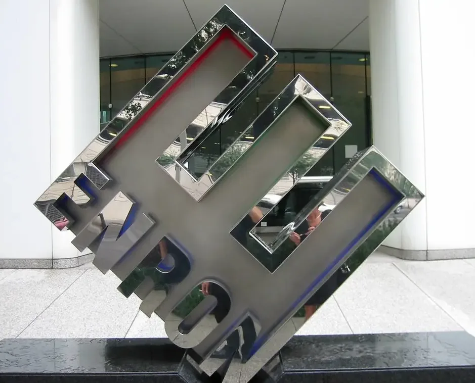 Photograph of a large tilted, mirrored Enron logo sculpture outside a glass office building entrance.