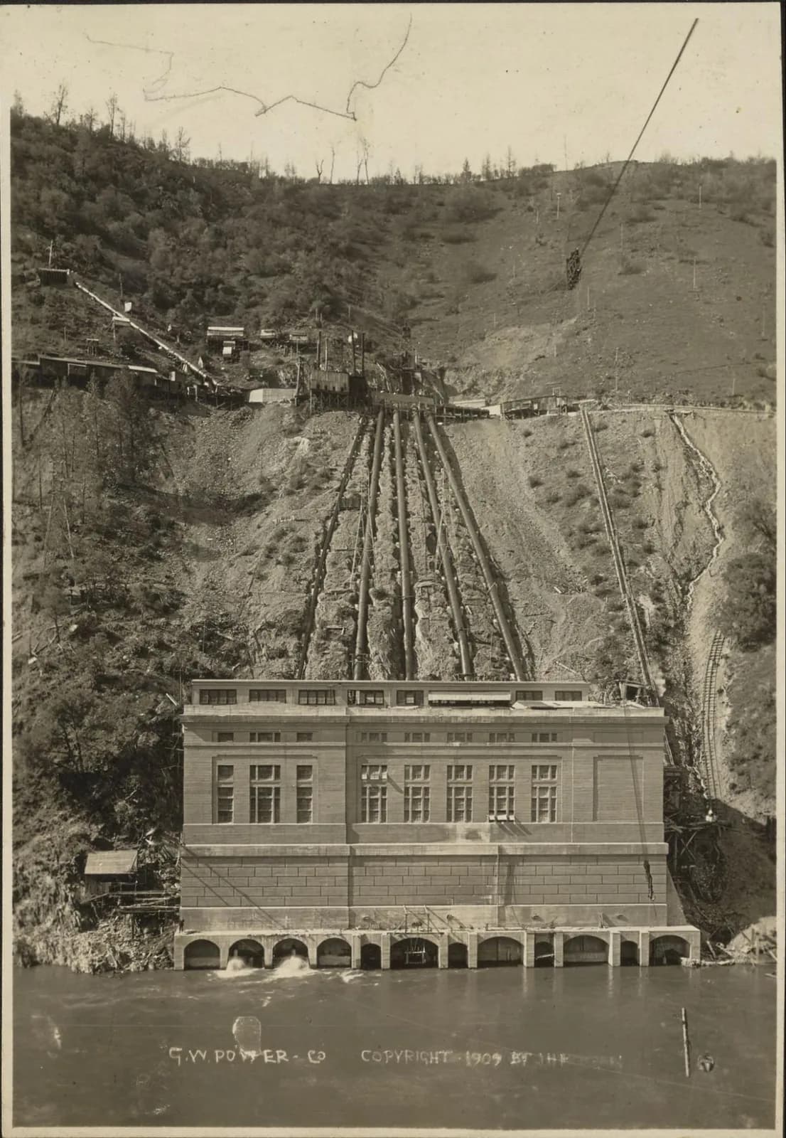 Black-and-white 1909 photograph of a riverside hydroelectric powerhouse with an inclined railway track running up a forested hillside behind it.